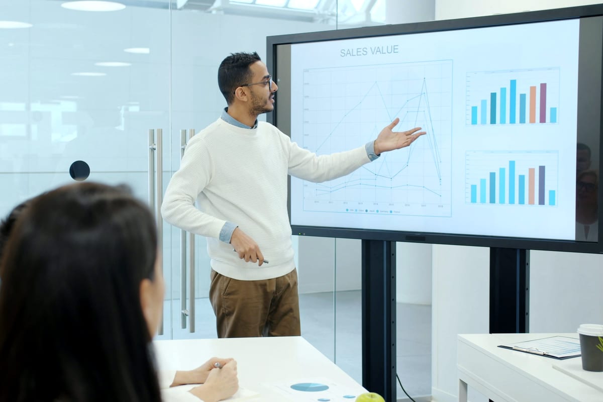 Trading mentor explaining market charts on a large screen to a small group of learners