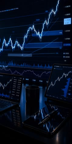 Dark trading desk with a laptop, tablet and phone showing blue price charts in front of a large chart screen